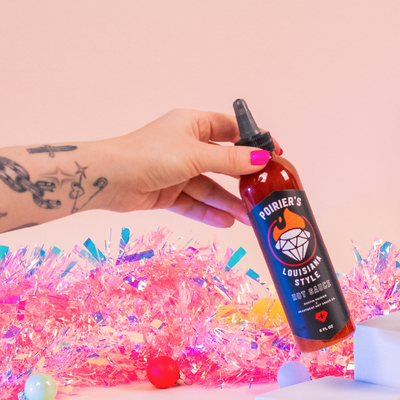 Hand holding a bottle of Poirier's Louisiana Style Hot Sauce with colorful confetti in the background