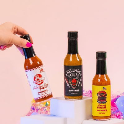 Hand holding a bottle of Hellfire Club hot sauce with other bottles on a festive background