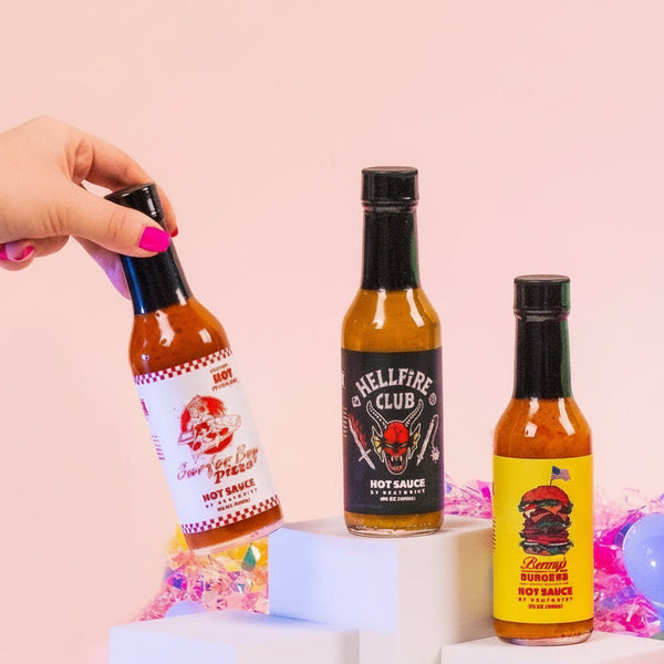 Hand holding a bottle of Hellfire Club hot sauce with other bottles on a festive background